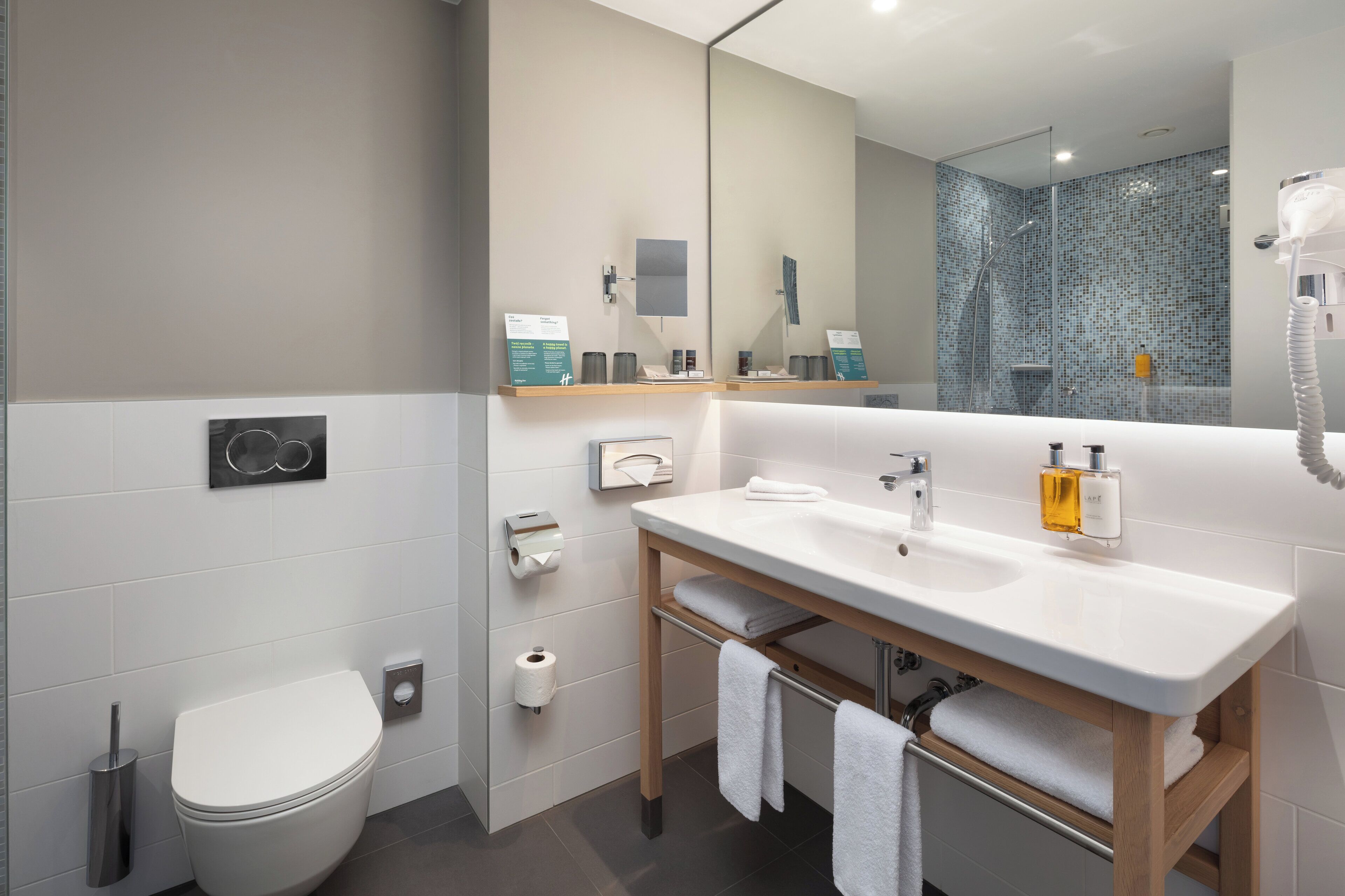 Een moderne badkamer met een wit wastafel, spiegel en toilet. Handdoeken hangen aan een rek en er is een douche in de achtergrond te zien.