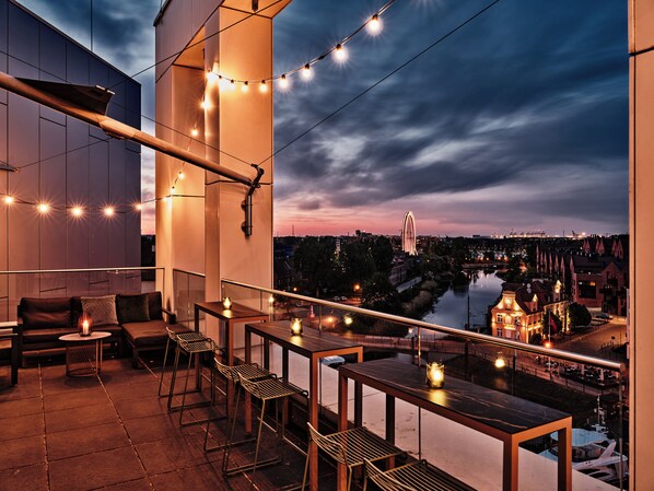 An outdoor terrace with a view of a river and cityscape, featuring a table and chairs, string lights, and a railing. - Holiday Inn Gdansk - City Centre by IHG (Gdansk)