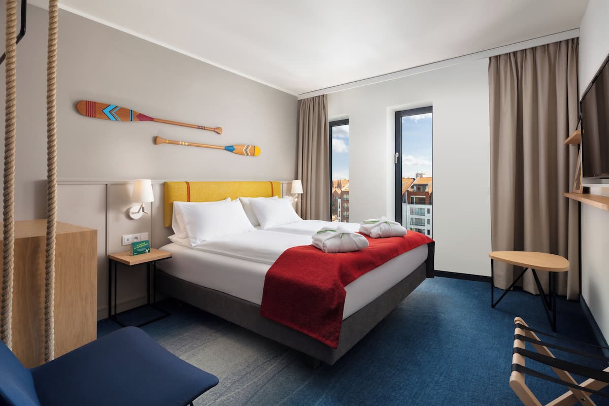 A hotel room with a large bed, a small table, a chair, a TV, and a window with a view of buildings.