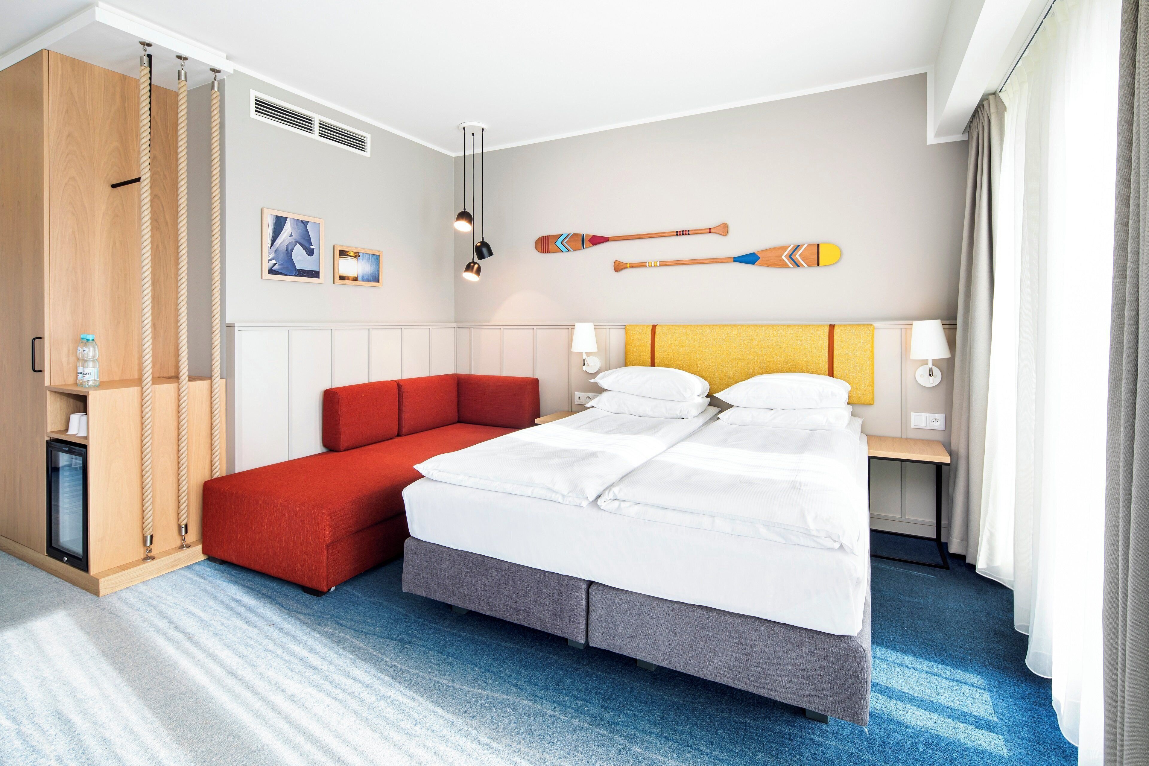 A hotel room with a bed, a red sofa, a wooden wardrobe, and two paddleboards on the wall.