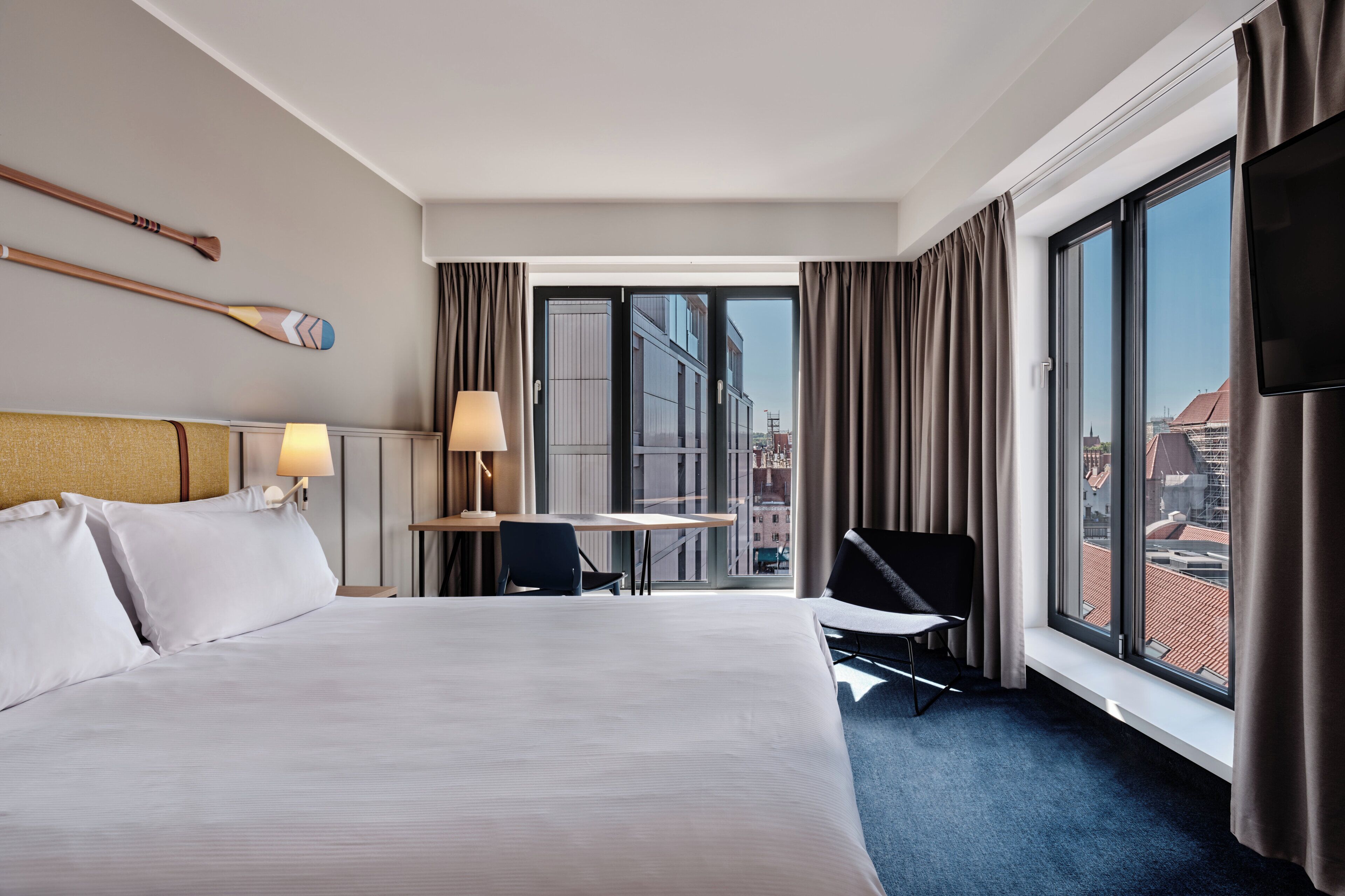 Une chambre d’hôtel avec un grand lit, un bureau avec une lampe, une chaise et une vue sur la ville grâce à de grandes fenêtres.