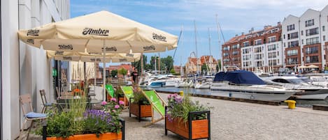 A waterfront café with outdoor seating, a large umbrella, and a view of boats and buildings.