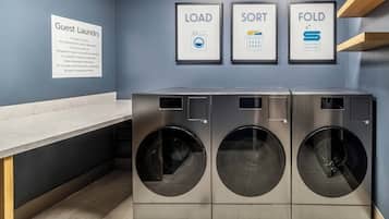 A laundry area with three stacked washing machines, a sign for guest laundry, and three framed instructions for loading, sorting, and folding.