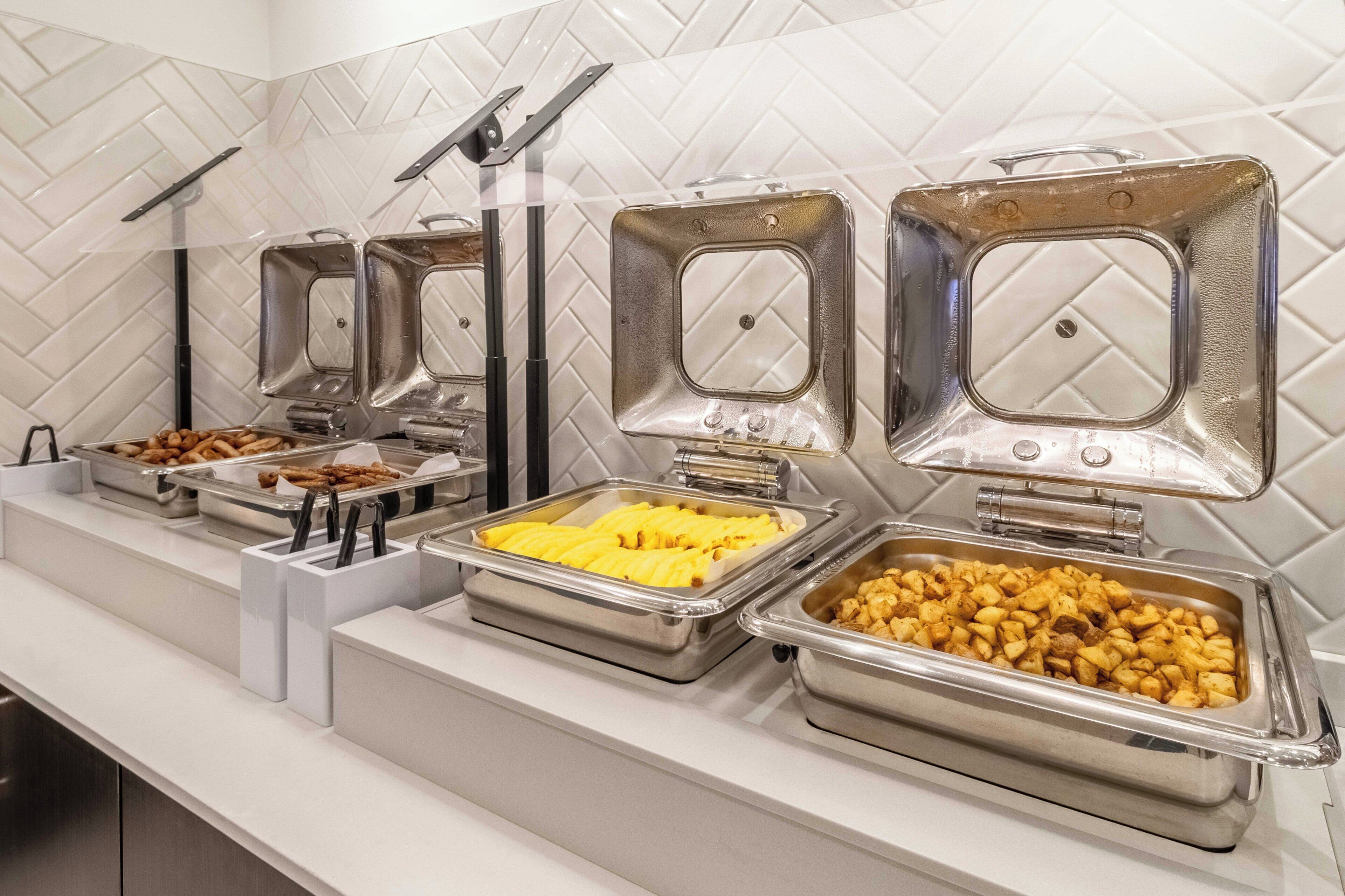 Un bufé con diversos alimentos en recipientes metálicos, dispuesto sobre una encimera blanca con salpicadero de azulejos.