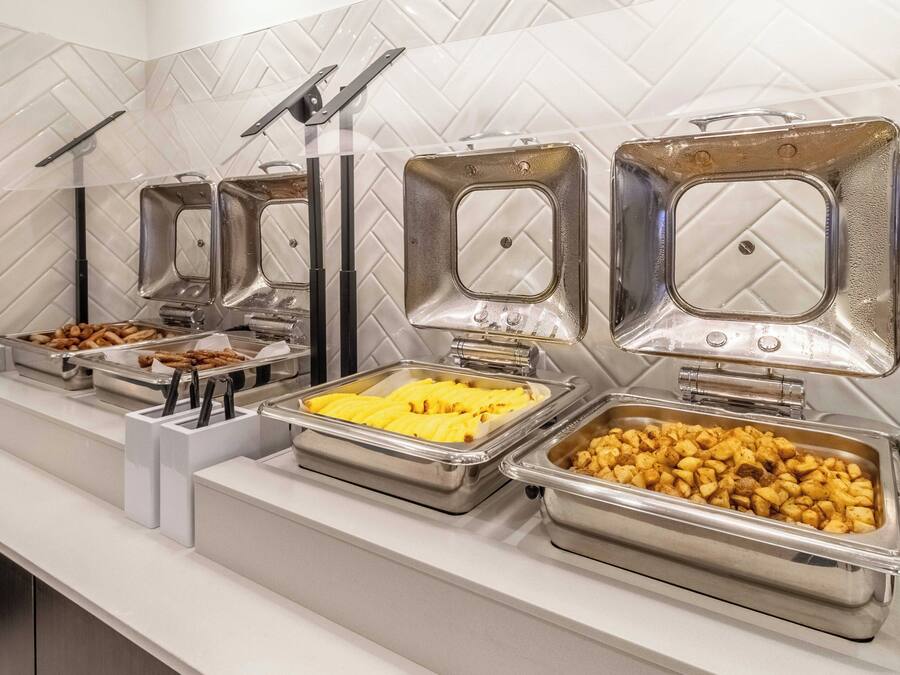 Un bufé con diversos alimentos en recipientes metálicos, dispuesto sobre una encimera blanca con salpicadero de azulejos.