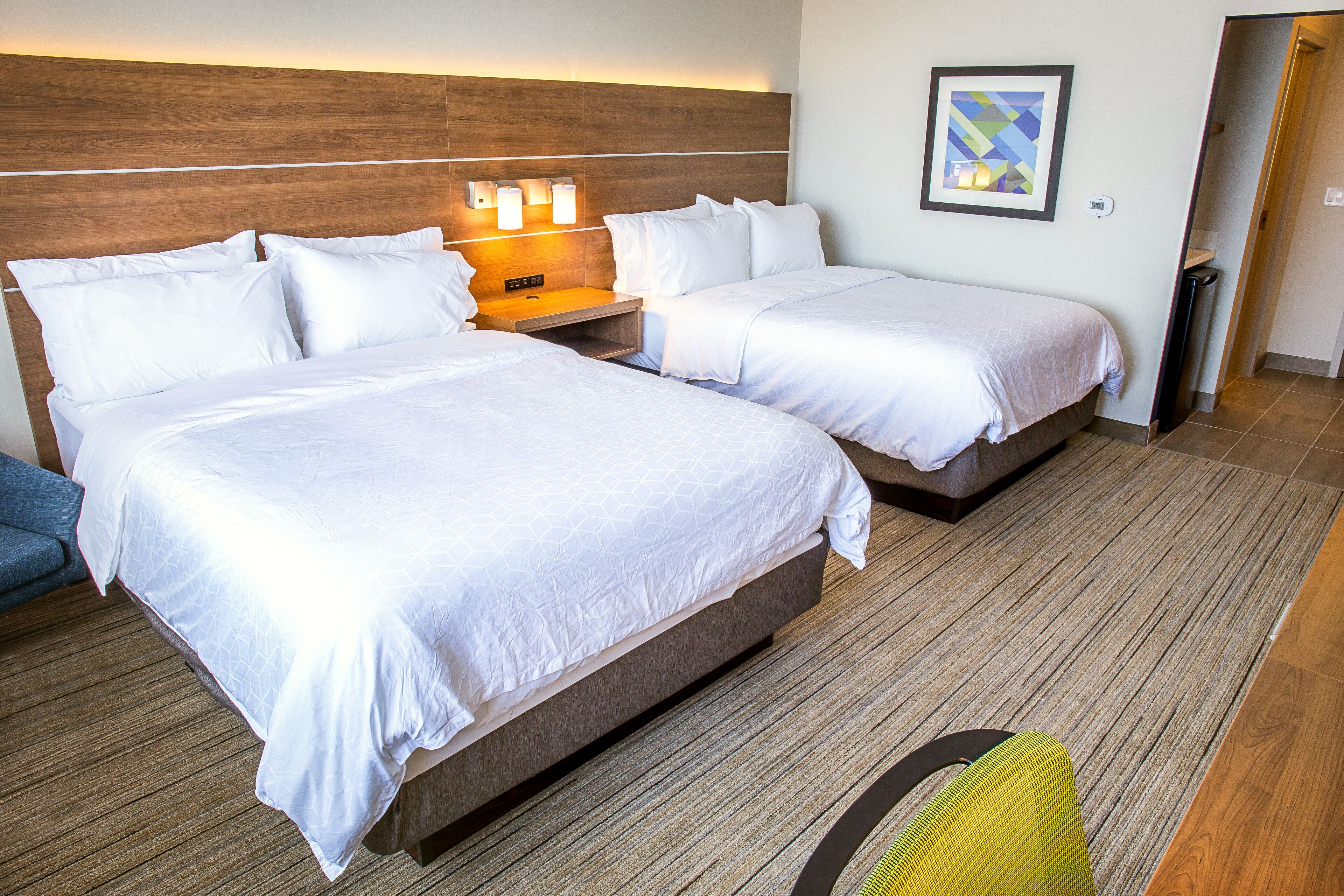 Habitación de hotel con dos camas, cabecero de madera, lámparas de noche, un cuadro en la pared y una silla.