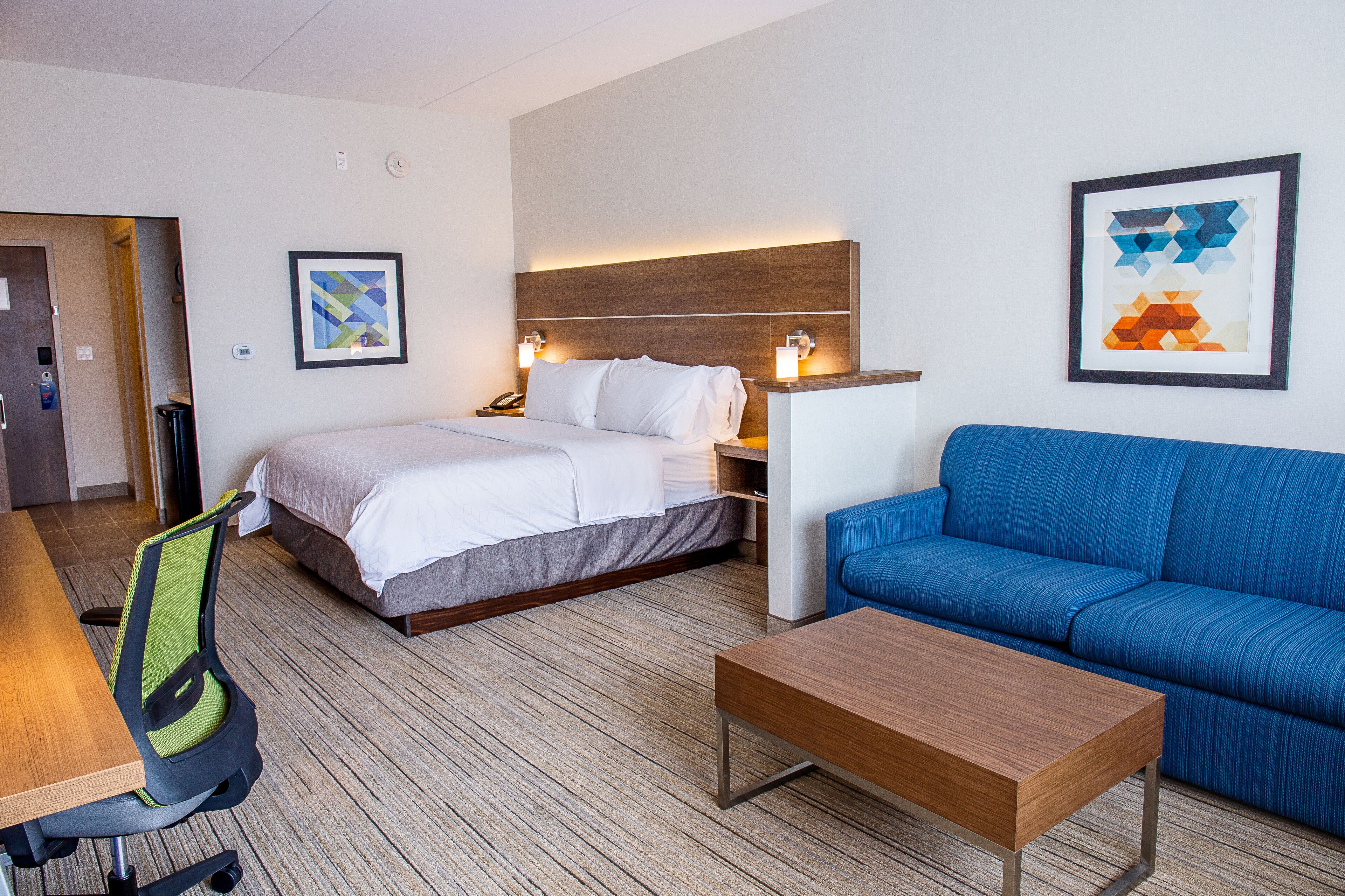 Ein Hotelzimmer mit einem Bett, einem blauen Sofa, einem Holztisch, einem Schreibtisch mit Stuhl und zwei gerahmten Kunstwerken an den Wänden.