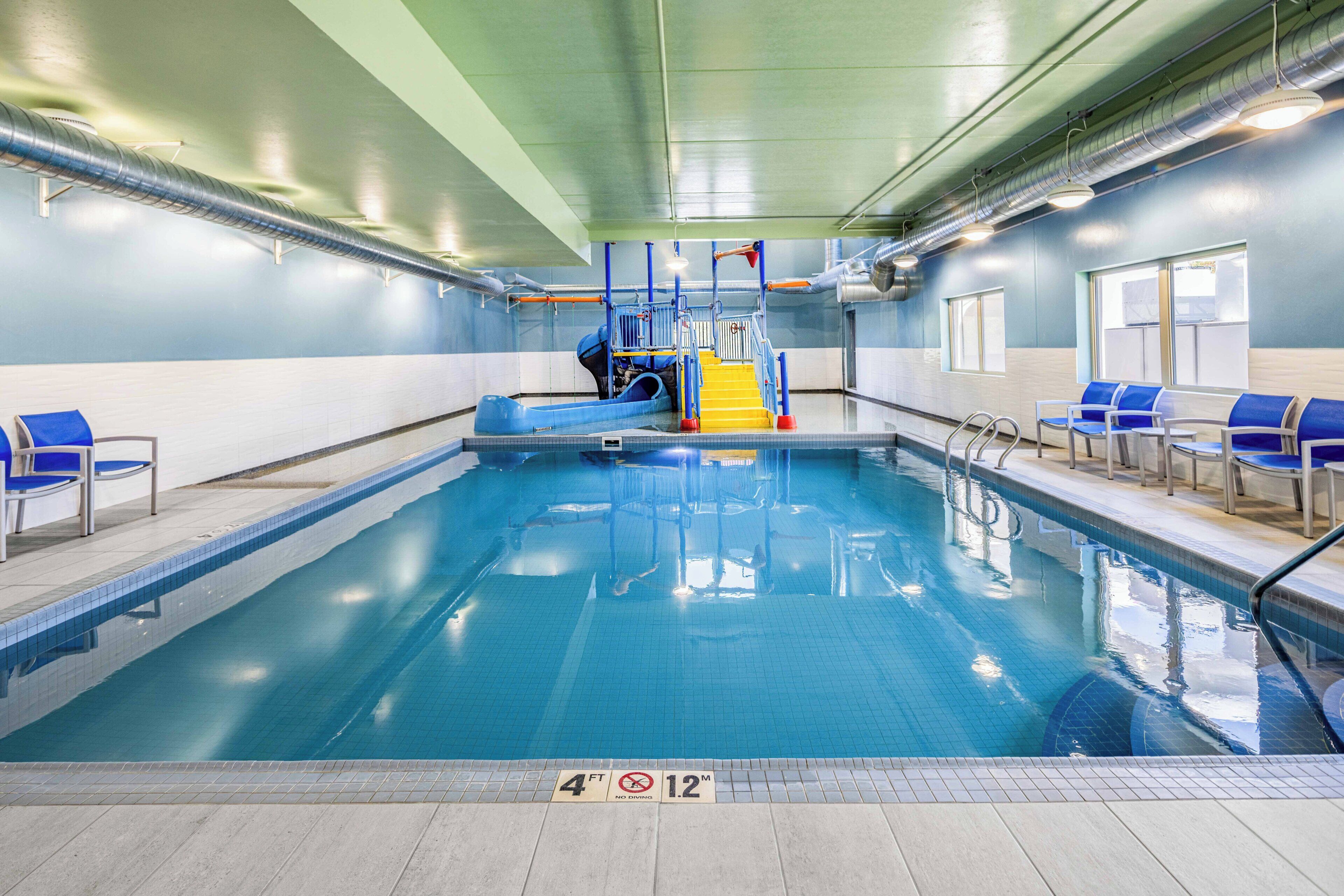An indoor swimming pool with a slide, blue chairs, and a lifeguard stand.