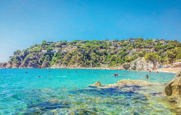 Ein Küstenstrand mit klarem türkisblauem Wasser, felsigem Ufer und einem Hügel mit Gebäuden.