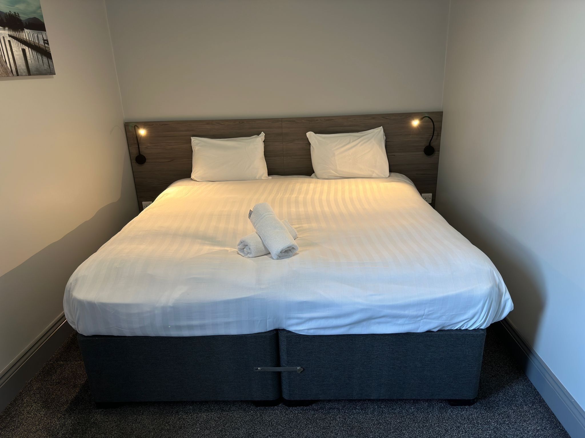 A neatly made bed with white linens and a folded towel, a wooden headboard, and a small wall-mounted light.