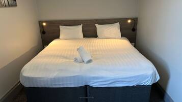 A neatly made bed with white linens and a folded towel, a wooden headboard, and a small wall-mounted light.