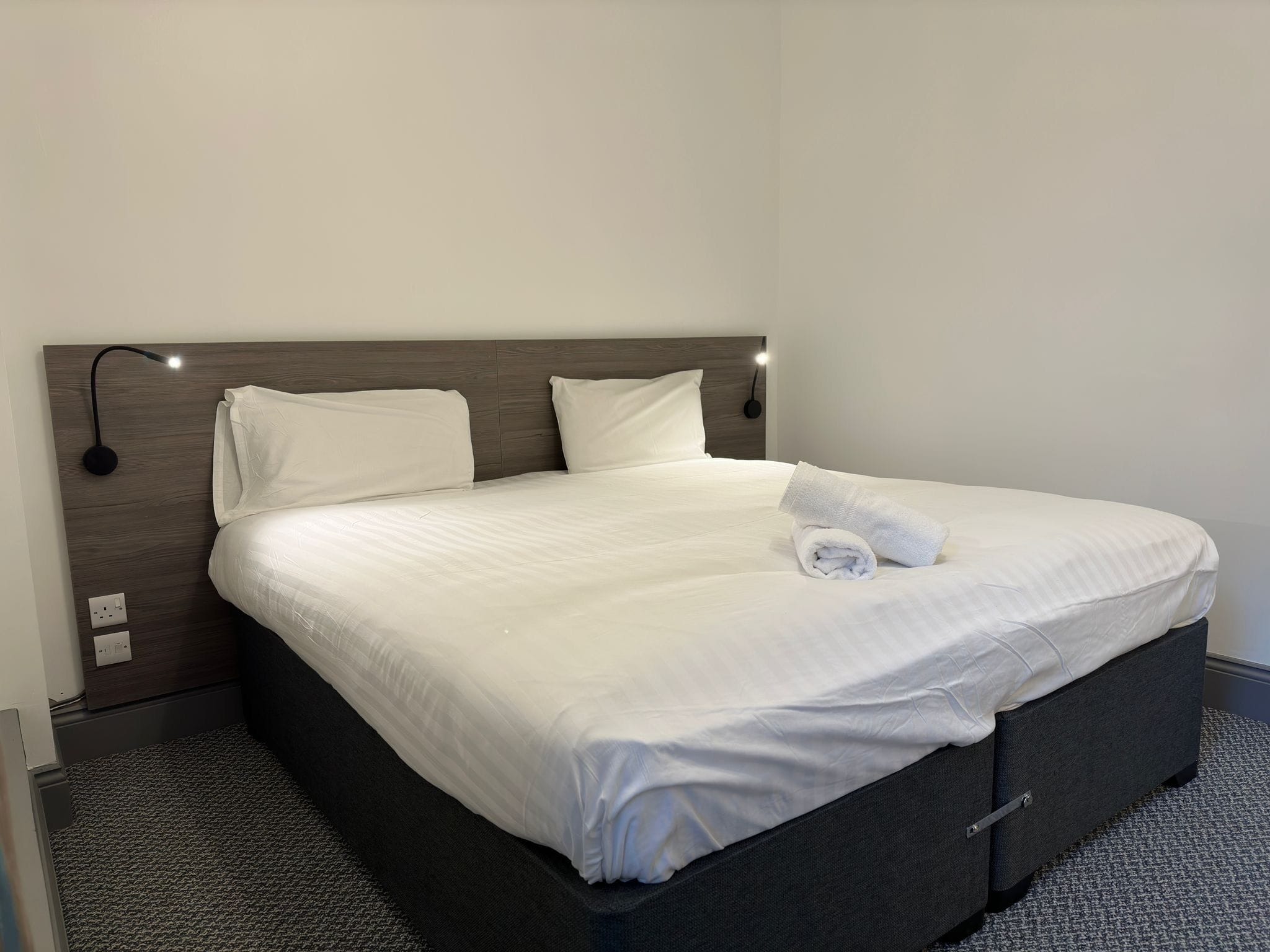 A neatly made bed with white linens and a rolled towel, a dark headboard, and a wall-mounted light fixture.
