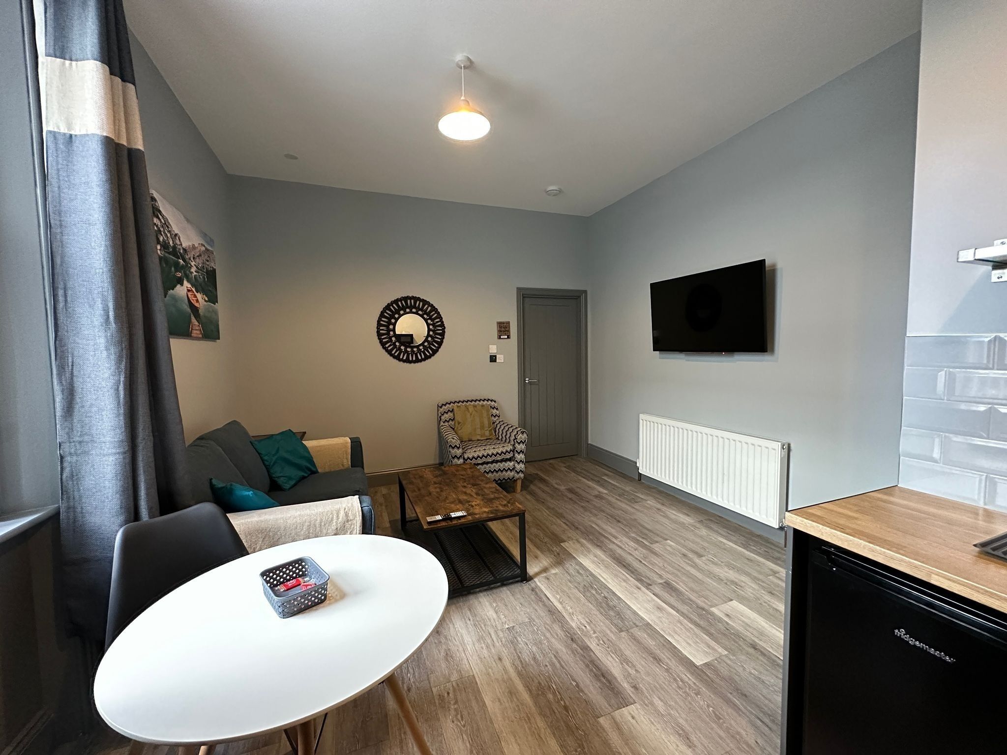 Ein modernes Wohnzimmer mit Sofa, Sessel, Couchtisch und Esstisch mit Korb. Ein Fernseher ist an der Wand montiert, und im Hintergrund ist eine Tür zu sehen.