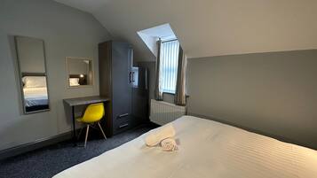 A hotel room with a bed, a desk, a chair, a wardrobe, a radiator, and a window with curtains.