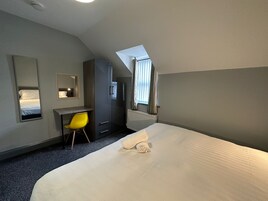 A hotel room with a bed, a desk, a chair, a wardrobe, a radiator, and a window with curtains.