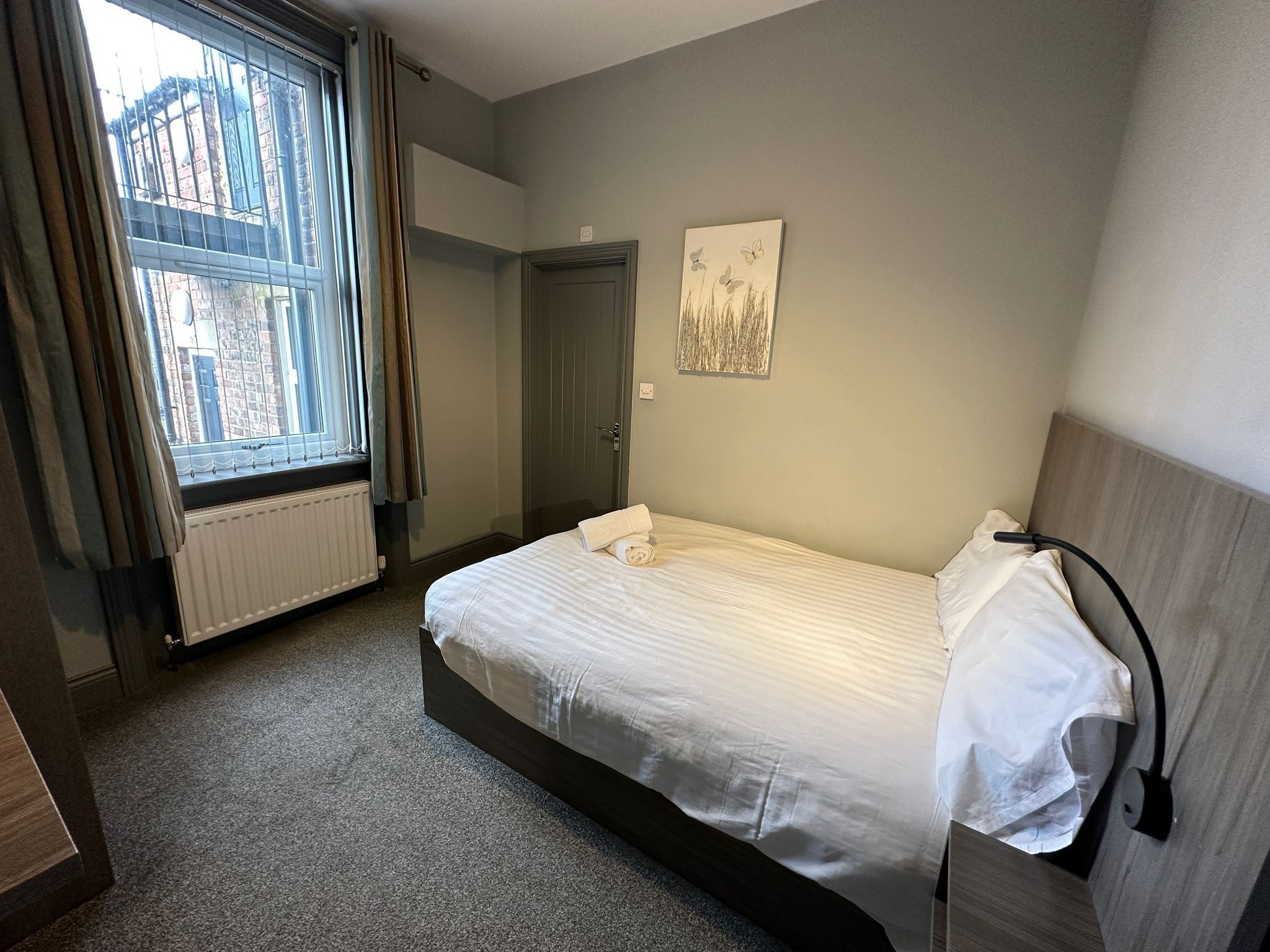 Een hotelkamer met een eenpersoonsbed, een radiator, een raam met gordijnen, een deur en een wandlamp.