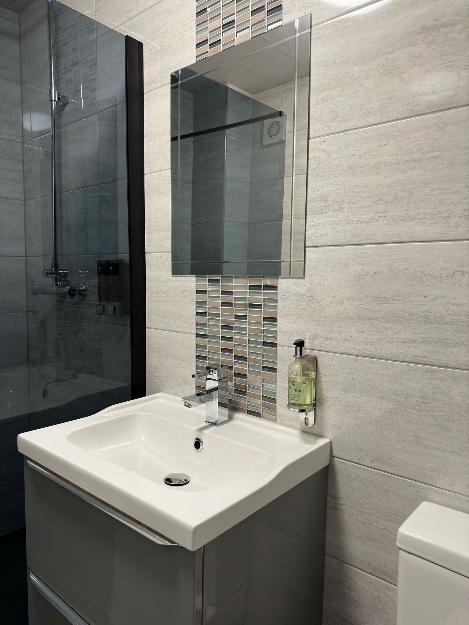 A bathroom with a glass shower enclosure, a white sink with a chrome faucet, a mirror above the sink, and a soap dispenser on the wall.