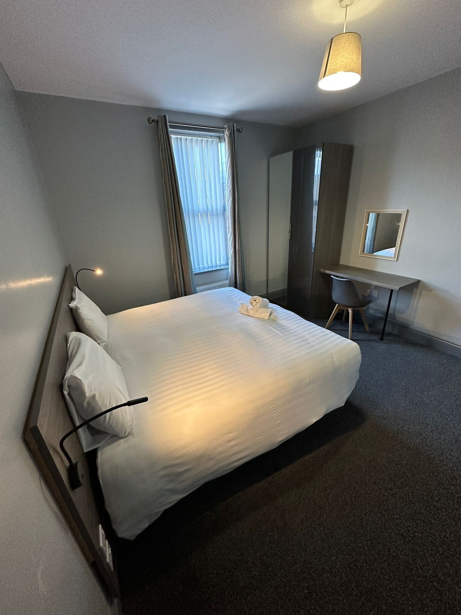 Una cama bien hecha con almohadas y una toalla doblada, un escritorio con una silla, un espejo y una lámpara colgante en una habitación de hotel.