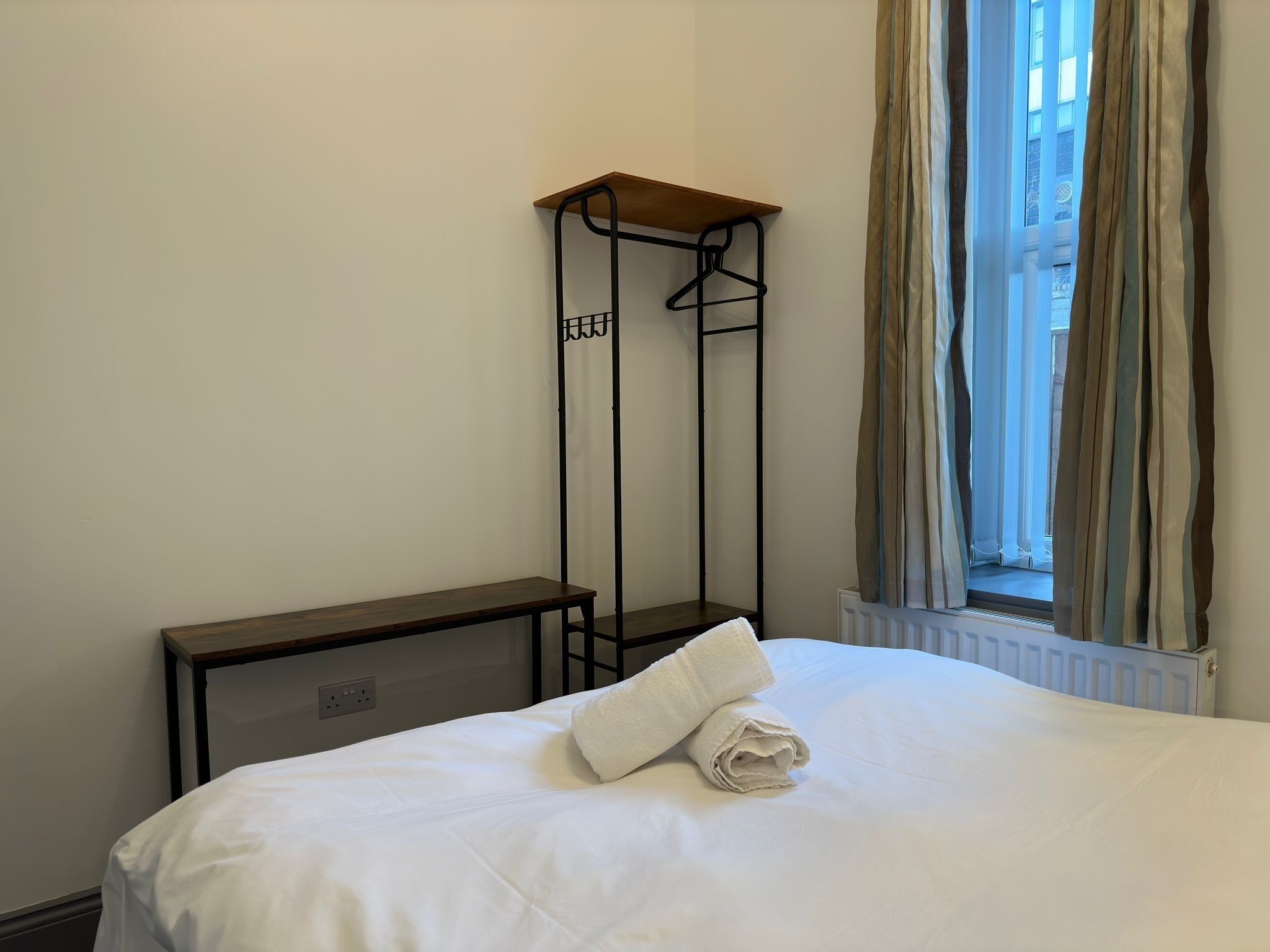 A hotel room with a bed, a bedside table, a hanging clothes rack, and a window with curtains.