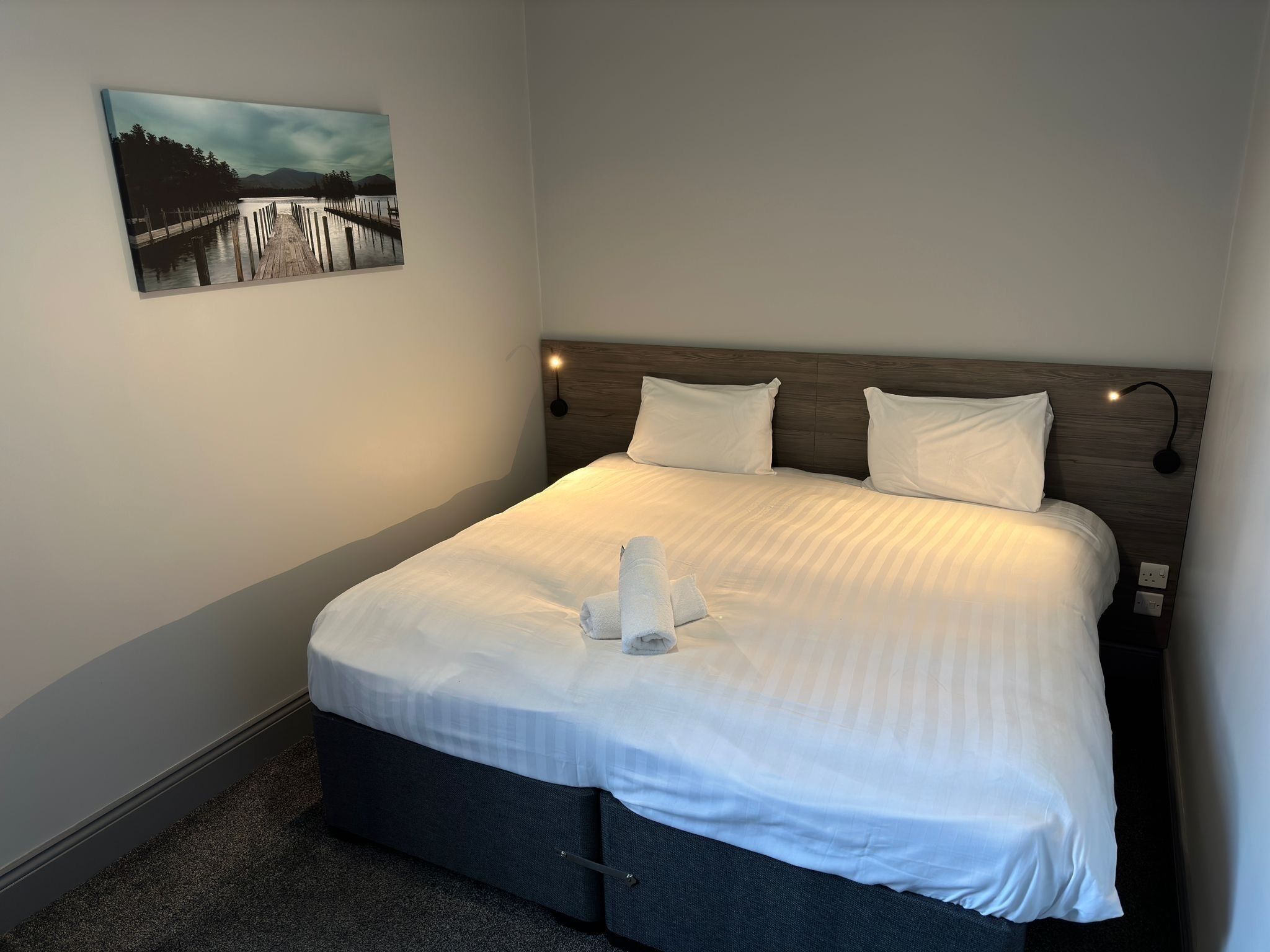 A neatly made bed with white linens, a headboard with wall-mounted lights, and a painting of a pier on the wall.