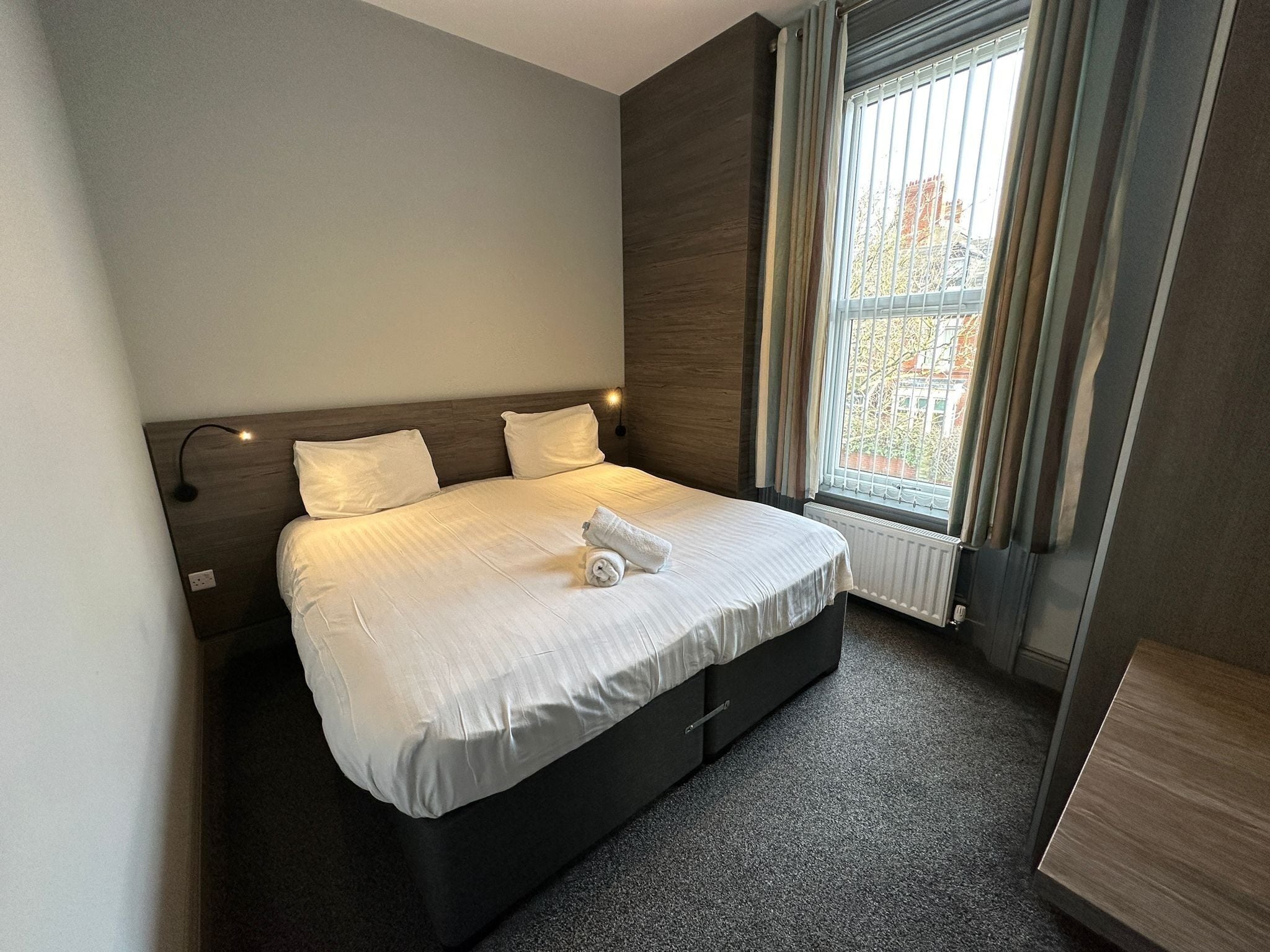 A modern hotel room with a large bed, a nightstand, a window with curtains, and a view of the outside.