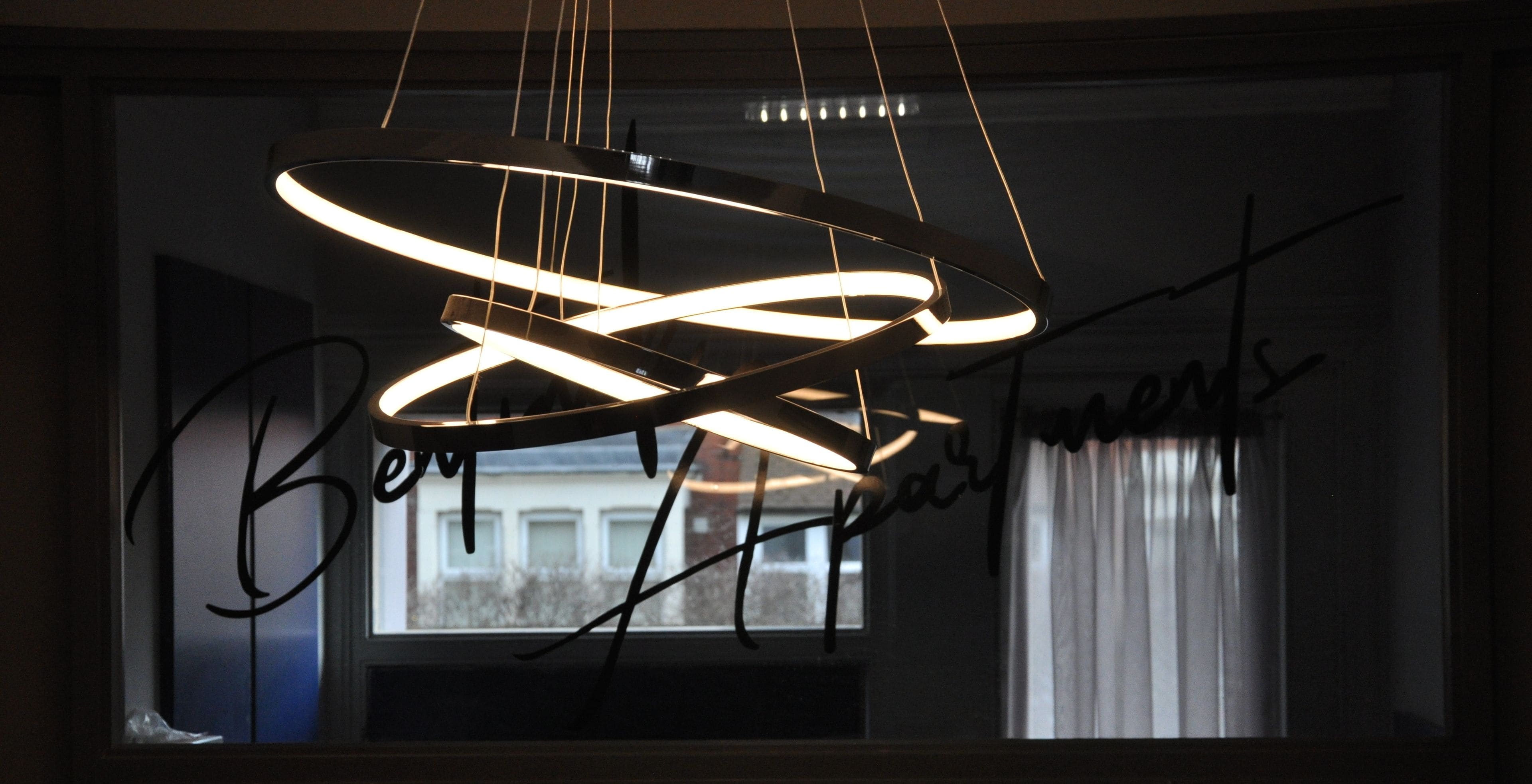A modern chandelier with a circular design, a window with a view of buildings, and a handwritten sign on the window.