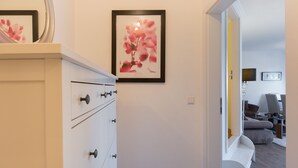 Un couloir avec une commode blanche, une œuvre d’art florale encadrée au mur, et un aperçu d’un salon avec un canapé et une table basse.