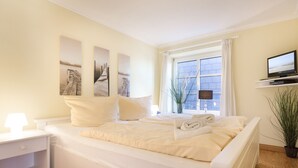 Une chambre à coucher avec un lit, une table de chevet, un téléviseur posé sur une étagère et une fenêtre avec des rideaux.