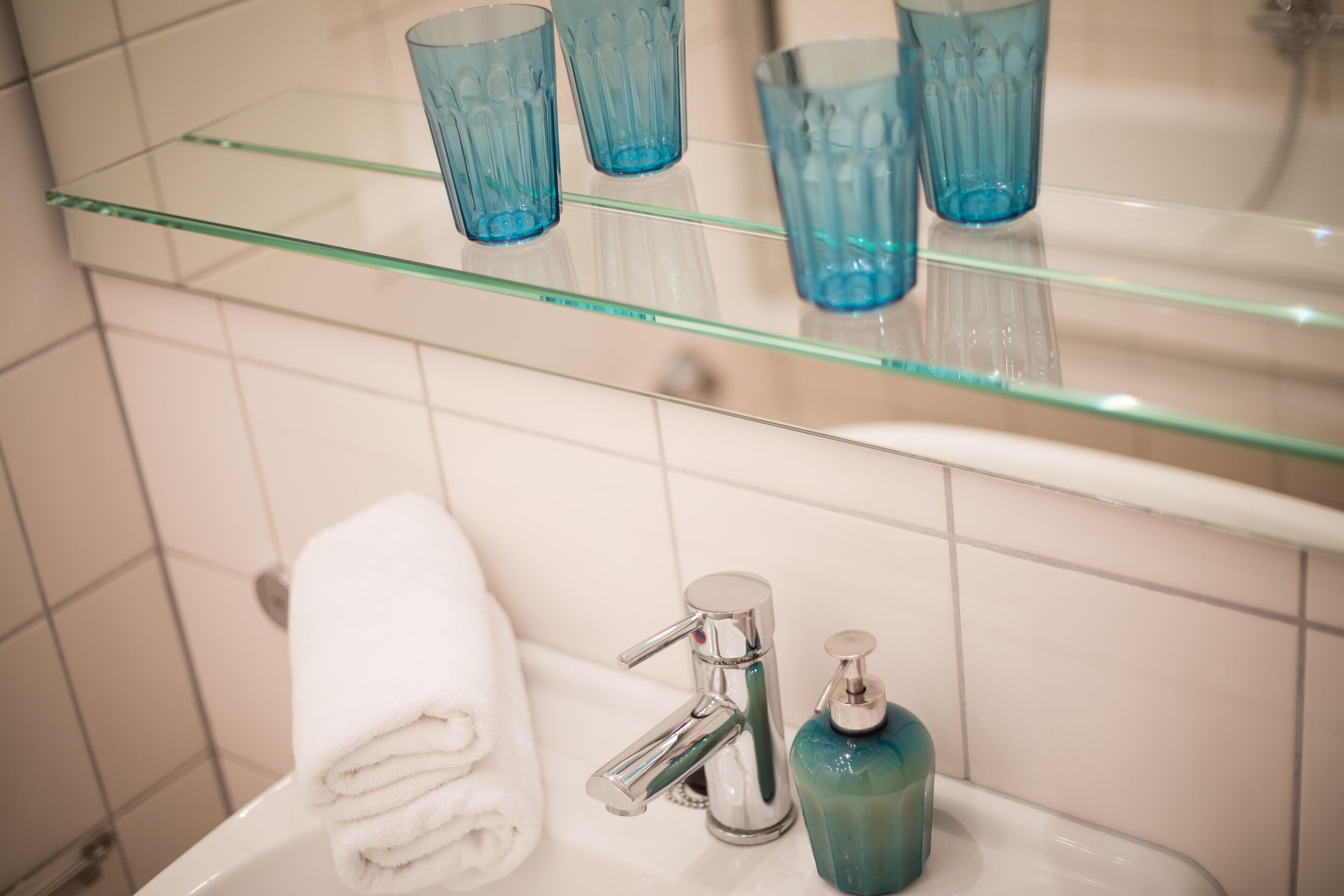 A bathroom with a glass shelf holding blue glasses, a white towel, a chrome faucet, and a soap dispenser.