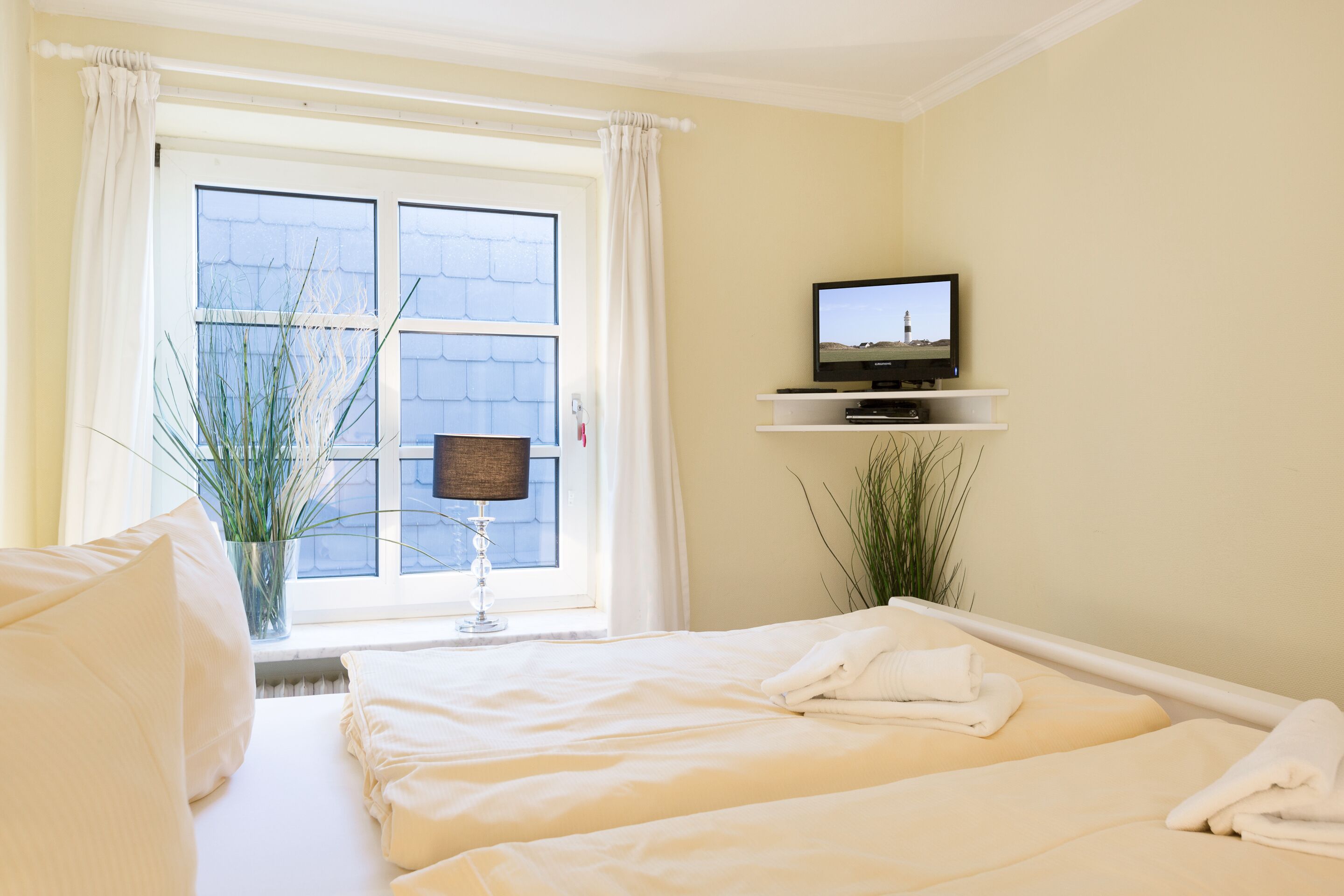 Una camera d'albergo con due letti, una televisione su un ripiano, una finestra con tende e una pianta.