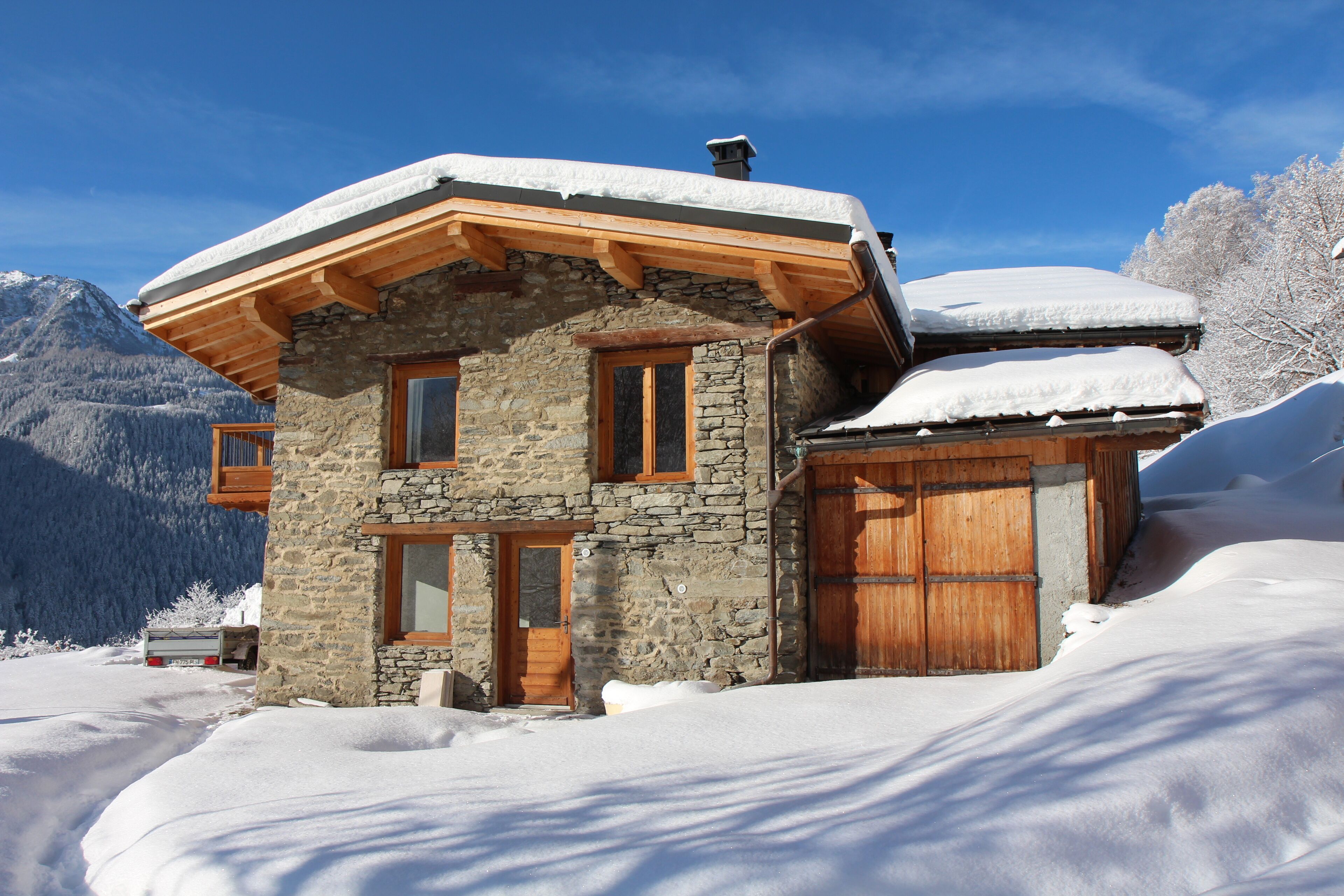 A stone building with wooden beams and a snow-covered roof, surrounded by snow and mountains.