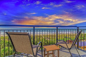Ein Balkon mit zwei Stühlen und einem kleinen Tisch, der Blick auf einen Strand unter blauem Himmel bietet.