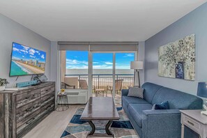 A living room with a TV, a sofa, a coffee table, and a view of the beach.