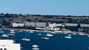 Un port de plaisance sur la côte, avec de nombreux bateaux amarrés, un grand bâtiment d’hôtel en arrière-plan et un paysage vallonné.