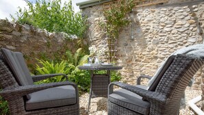 En terrasse med fletstole og et bord, omgivet af stenvægge og grøn vegetation.