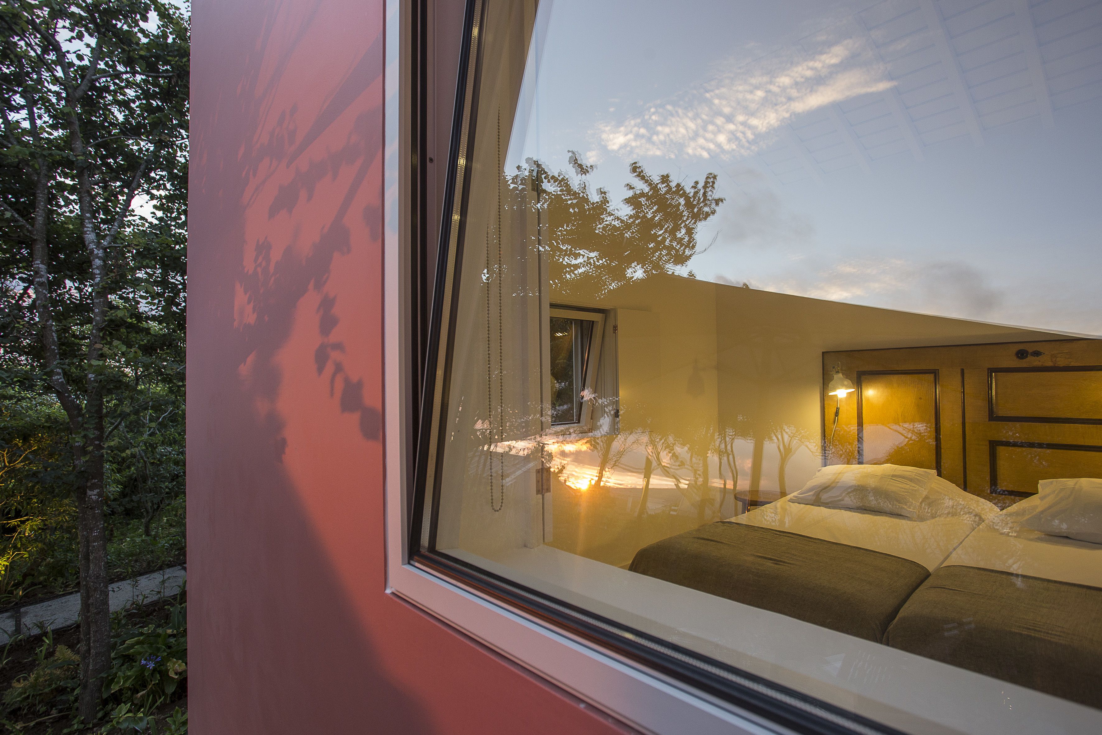 A bedroom with a large window, a bed with pillows, and a nightstand with a lamp.