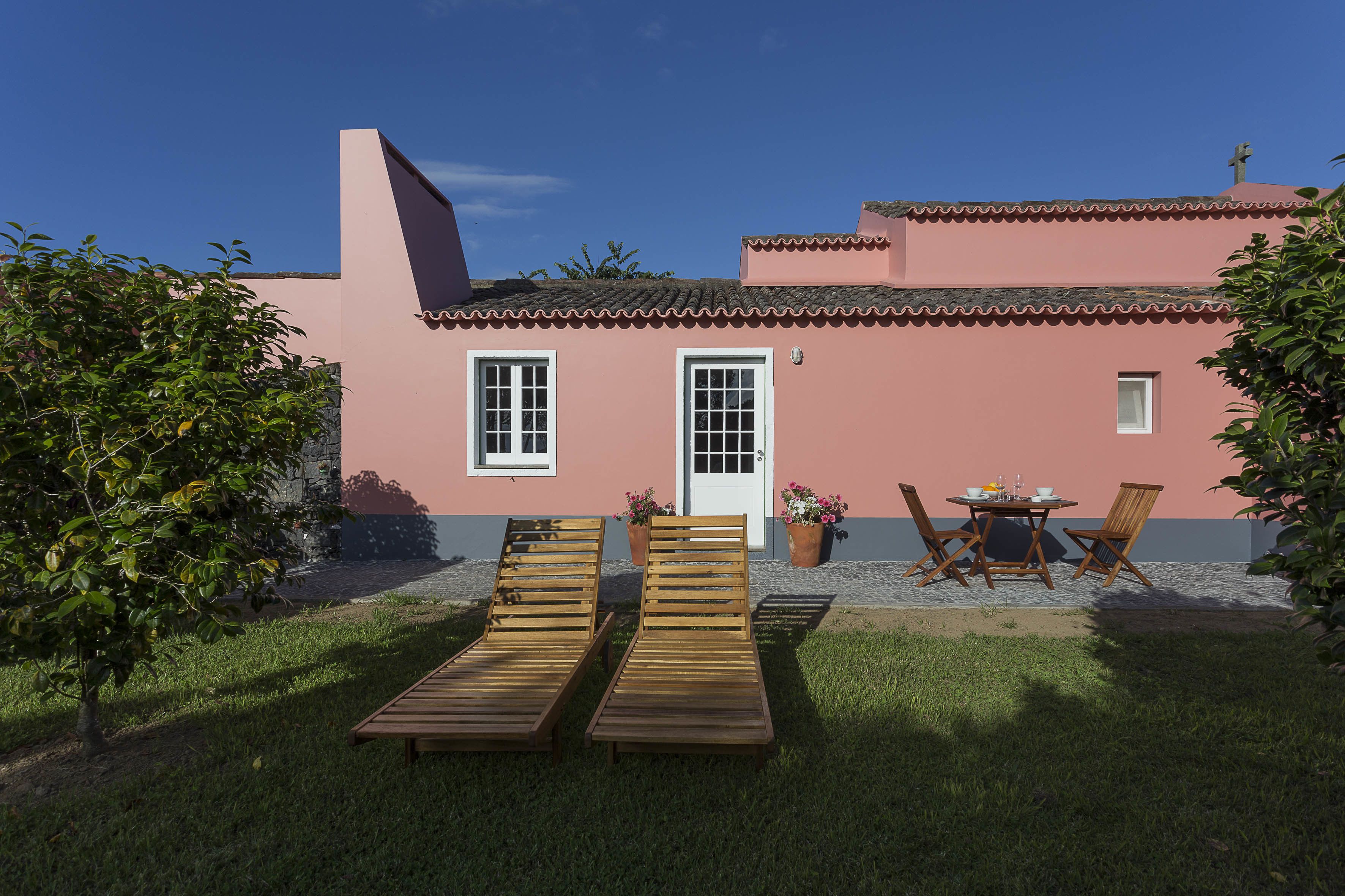 A pink building with a tiled roof, a white door, and a small window. There are two wooden lounge chairs and a table with chairs in the front yard.