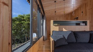 Eine moderne Holzhütte mit Holzvertäfelung, einem großen Fenster mit Blick auf Grünflächen, einer Couch mit Kissen und einem Regal mit einem Radio.