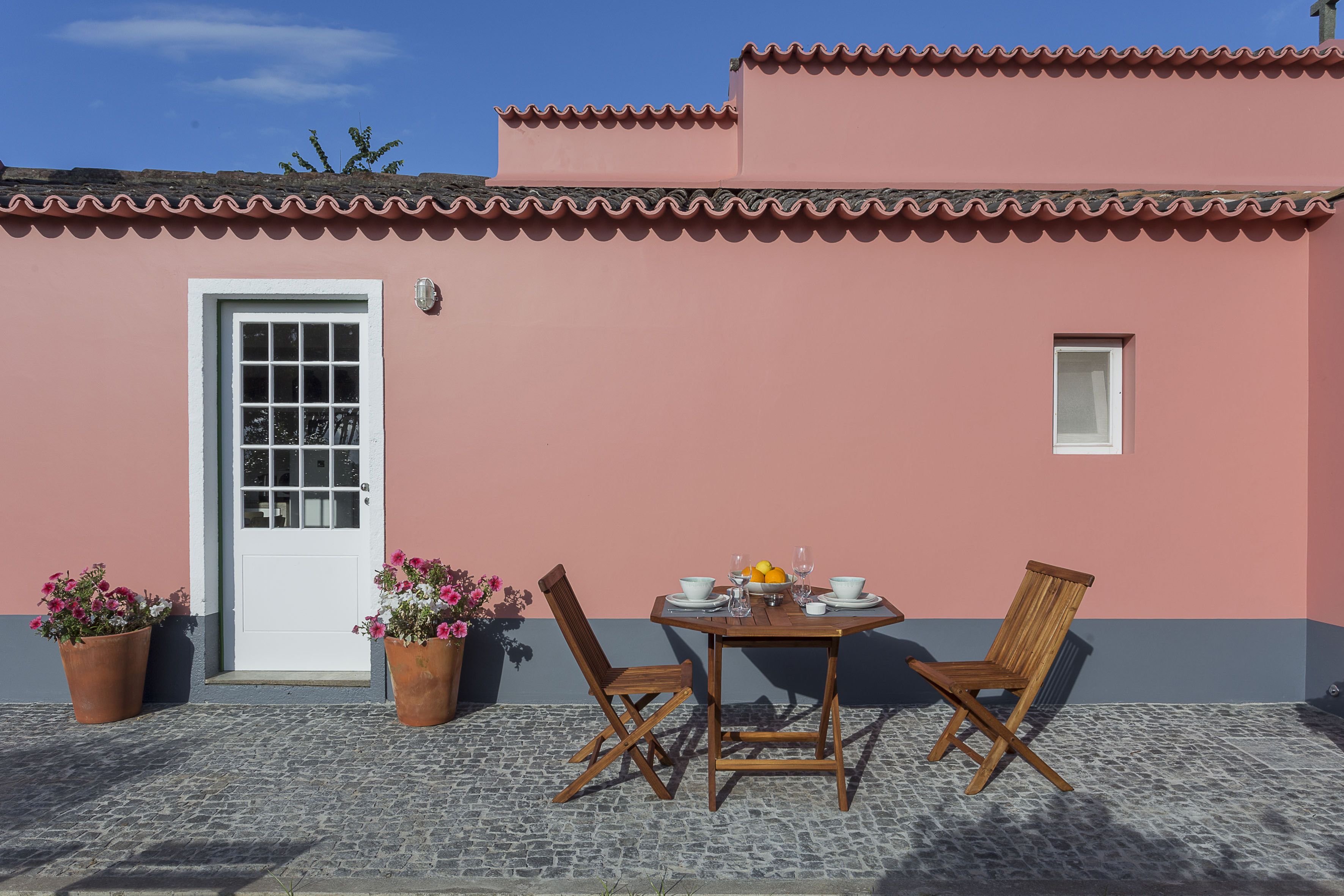 Un bâtiment rose avec un toit en tuiles, une porte blanche et une petite fenêtre. On y trouve une table avec des chaises et un panier de fruits.