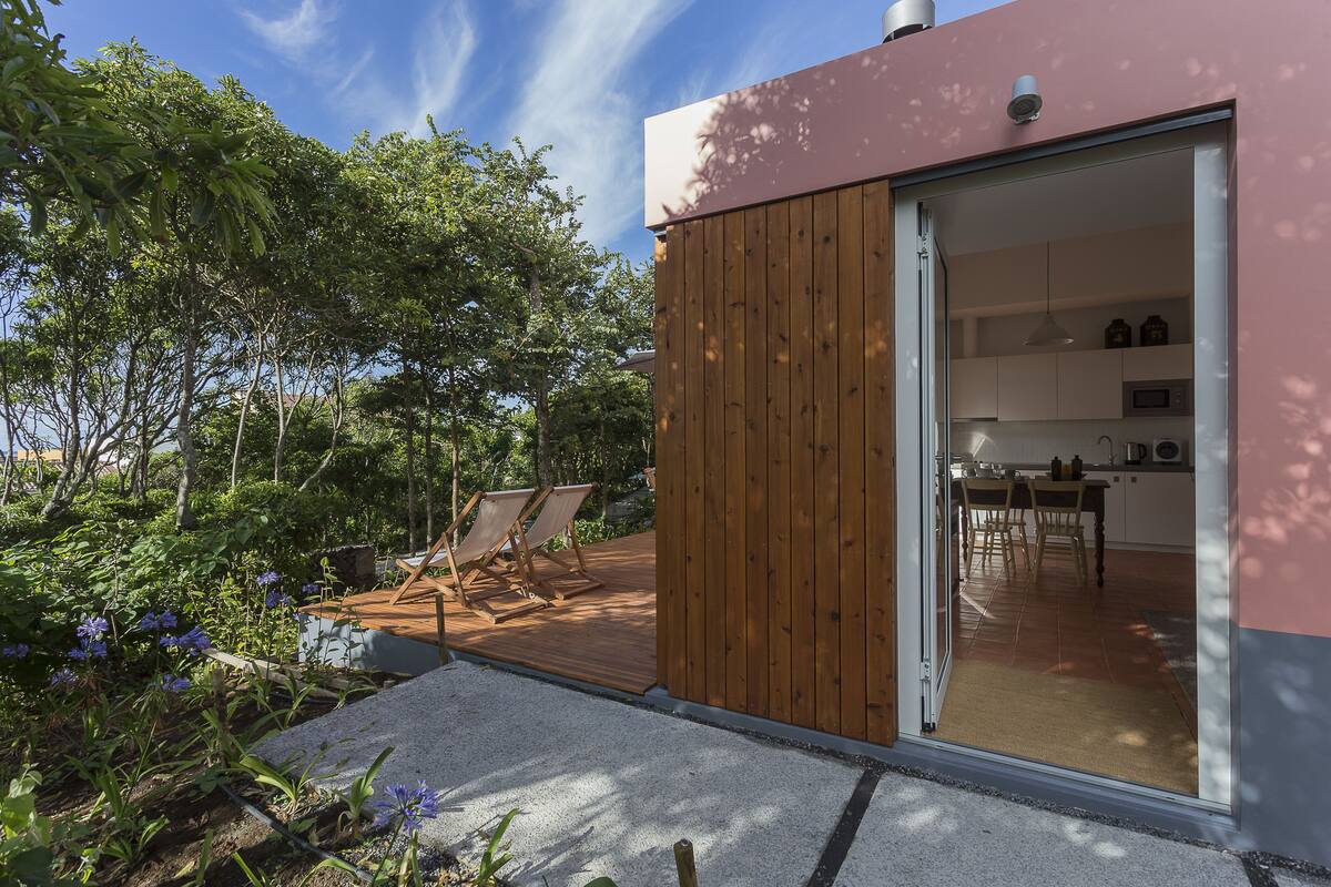 Une maison moderne dotée d’une terrasse en bois, d’une porte-fenêtre coulissante donnant sur la cuisine et d’un jardin agrémenté de plantes et d’un patio.