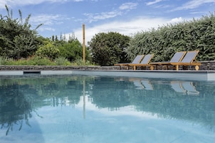 Una piscina con agua cristalina, rodeada de follaje y sillones de madera.