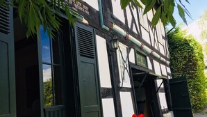 A traditional half-timbered building with a green door and windows, a stone wall, and red flowers in a pot. - Musikhaus Medebach. This historic house is in the village in Hochsauerland. (Medebach)