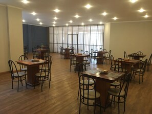 A spacious dining area with multiple wooden tables and chairs, large windows with curtains, and a high ceiling with recessed lighting.