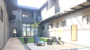 Un área de piscina exterior moderna con sillones de descanso, un pequeño jardín y un edificio con grandes ventanales.