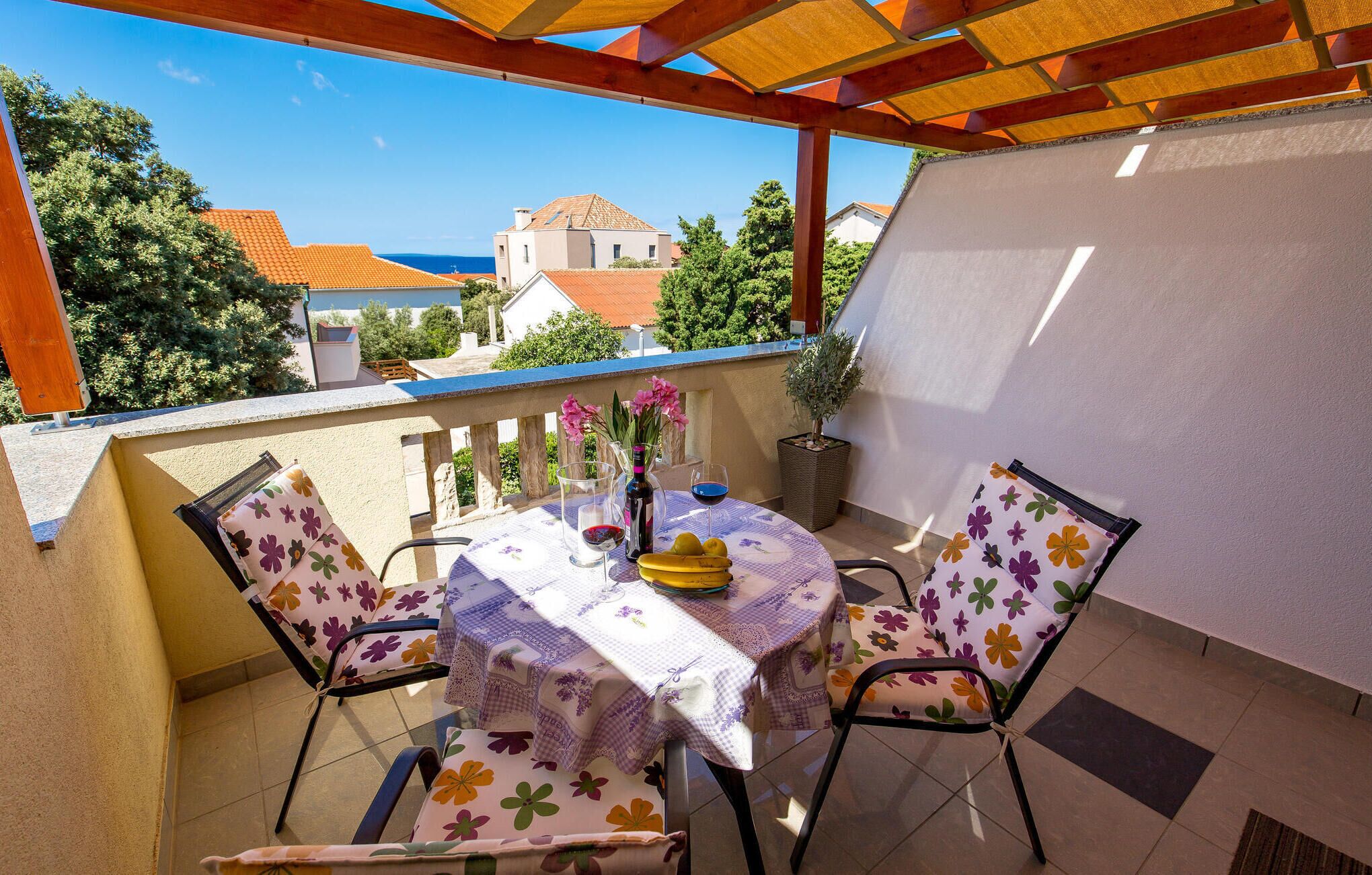 Ein überdachter Außenbereich mit Tisch und Stühlen, einem Blumentopf und Blick auf das Meer sowie Gebäude im Hintergrund.
