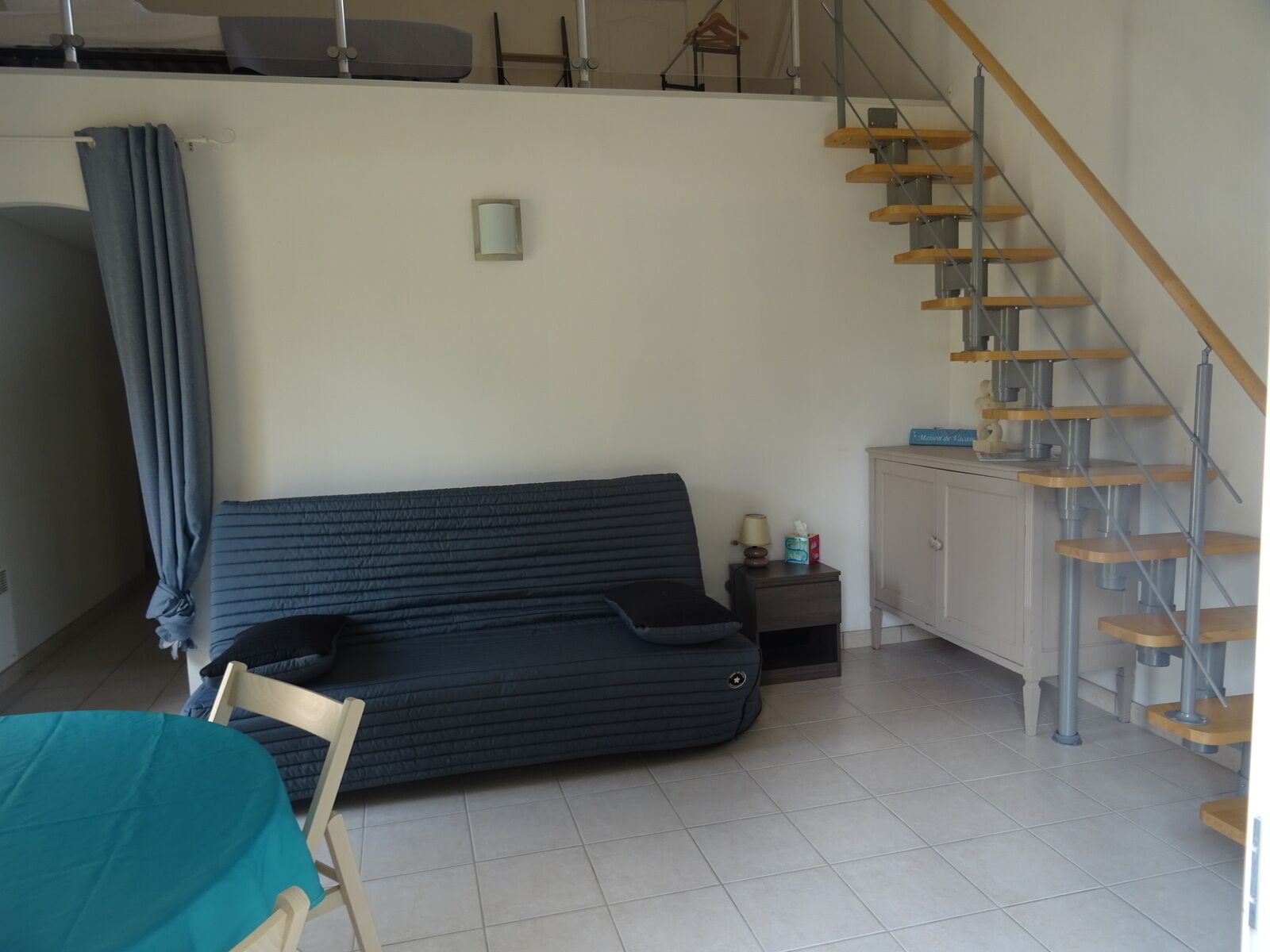 Una sala de estar con un sofá azul oscuro, una silla de madera, una pequeña mesa con mantel turquesa, un mueble aparador y una escalera de madera.