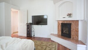 A bedroom with a bed, a television mounted on the wall, a fireplace, and a door leading to another room. - Easy Bay Hidden Gem (Richmond)