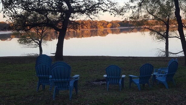 Un entorno junto al lago con cuatro sillas Adirondack dispuestas en semicírculo frente a un lago tranquilo, rodeado de árboles y césped.