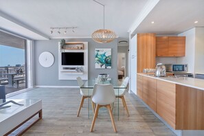 A modern kitchen with wooden cabinets, a glass dining table, and a built-in TV. - Horizon Bay 903 Fresh Beachfront Hideaway (Bloubergstrand)