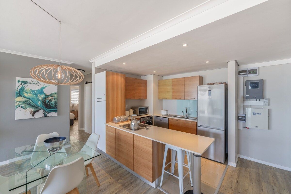 A modern kitchen with wooden cabinets, a stainless steel refrigerator, and a glass dining table with chairs.